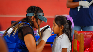 A woman wearing medical examination glasses shines a torch into a young girl's eyes to check for trachoma.