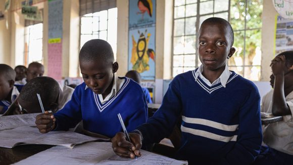 Two students wearing royal blue school jumpers sit at a desk in a classroom writing in exercise books.