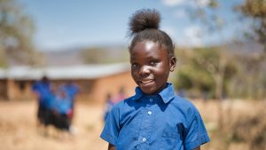 A girl wearing a blue shirt smiles while standing in a school playground.