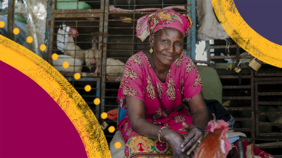 A woman wearing a pink patterned headwrap and gold earrings. Behind her, there are chickens in a cage.