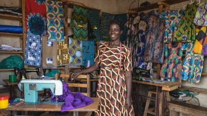 A woman stands next to a sewing machine in a workshop with colour fabric displayed on the walls. She has a tape measure around her neck.