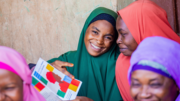 Four women wearing colourful head scarves look very happy. One woman holds a family planning leaflet.