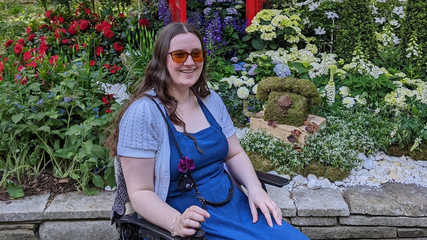 A woman with long brown hair sits in an electric wheelchair. Behind her is a curated floral display with colourful flowers and green foliage.