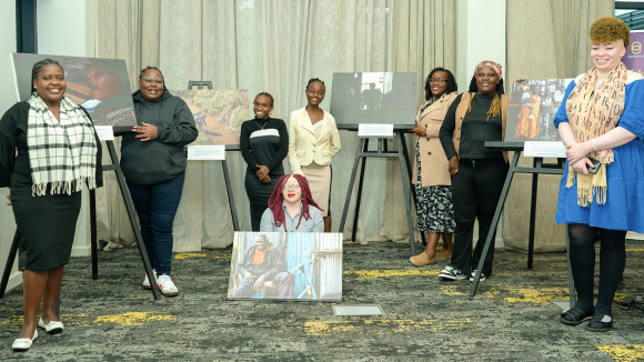Eight women proudly smile beside large canvases on easels.