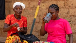 A woman drinks from a cup after taking medication. A female community volunteer sits next to her holding a pill bottle.