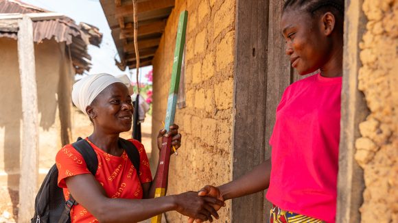 A female community volunteer carrying a dose pole shakes the hand of a woman standing in a doorway.