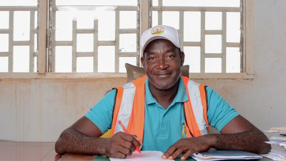 A man wearing a baseball cap and high-vis vest sits at a table writing.