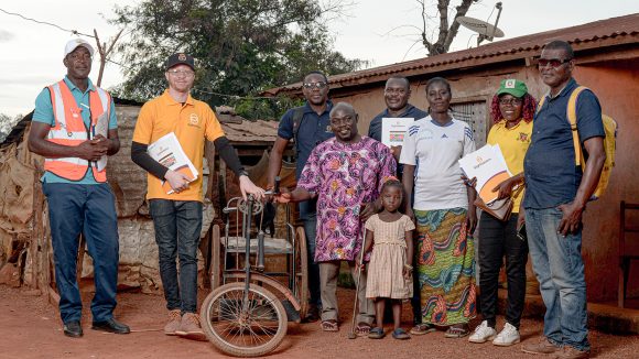 A group of men and women stand outside a house. One of the men is standing next to a mobility tricycle, while another man wearing a Sightsavers T-shirt holds paperwork.