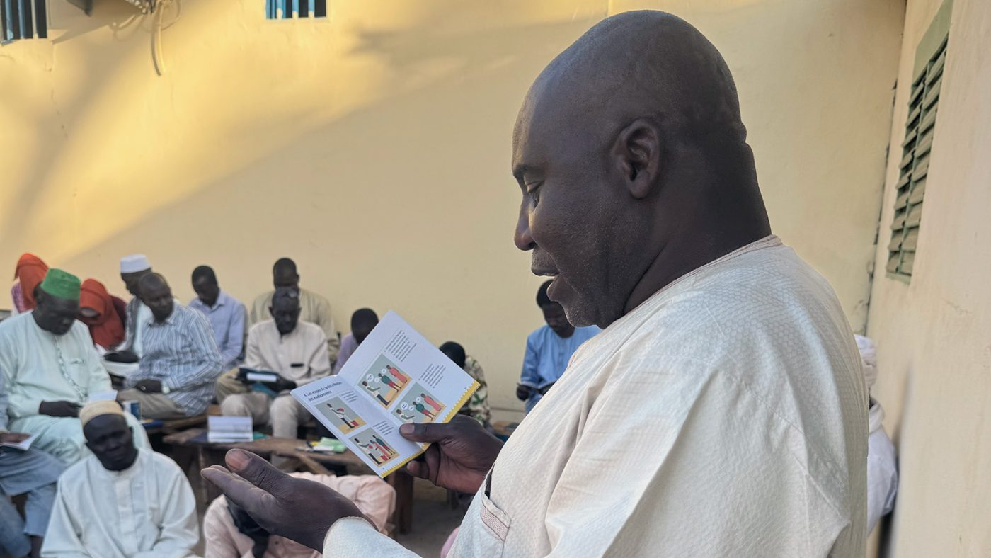 A man reads from a passport-sized document while a male audience listens. Inside, there are colourful illustrations and guidance.