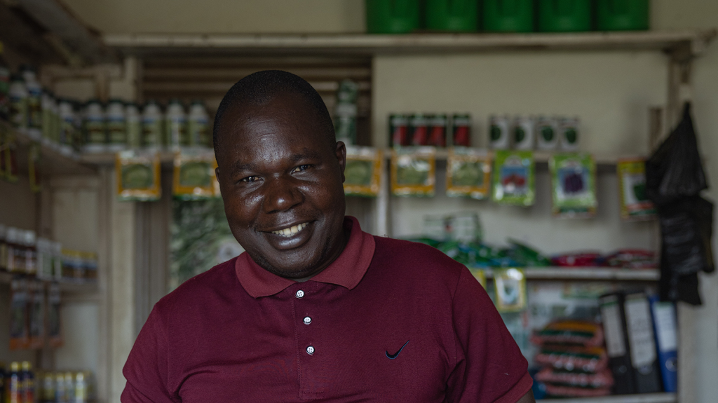 Denis smiles while standing in his shop. Behind him are farming supplies.