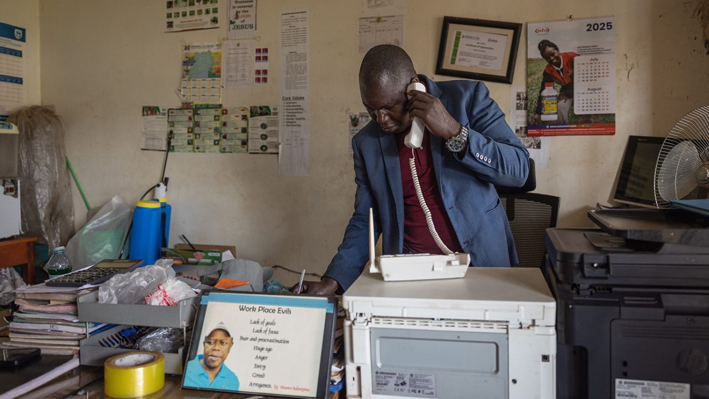 Denis stands in an office holding a phone to his ear.