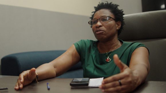 A woman sits at a desk talking to another person.
