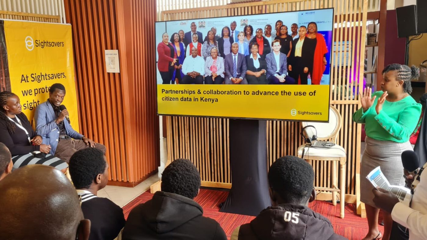 An audience watches a presentation. To the right of the stage, a woman is translating using sign language.