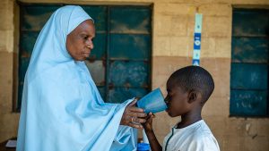 A boy drinks from a cup of water, held by an older woman, after taking medication to treat disease.