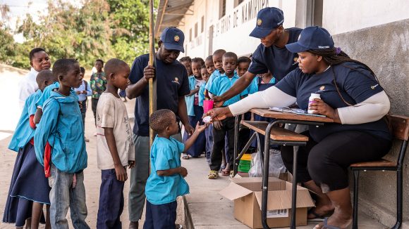 Students queue to receive medication. A female volunteer sitting at a desk hands a young child a tablet.