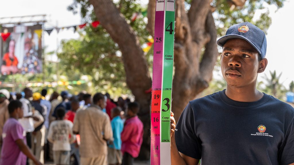 Momade, wearing branded clothing from the Ministry of Health, holds a dose pole.