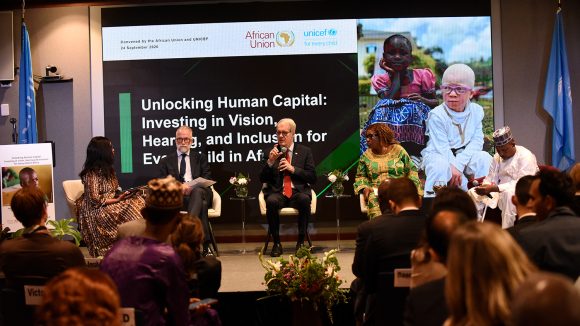 Five men and women sit on a stage during an event. A man in the centre is speaking into a microphone.